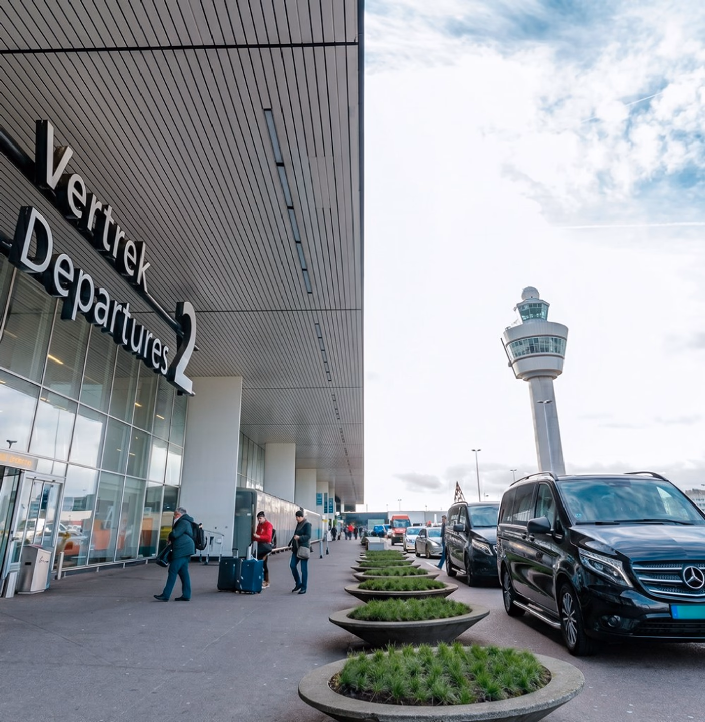 Black Mercedes-Benz chauffeur vans outside Amsterdam Airport Schiphol (AMS) Departures terminal with the control tower for a private airport transfer