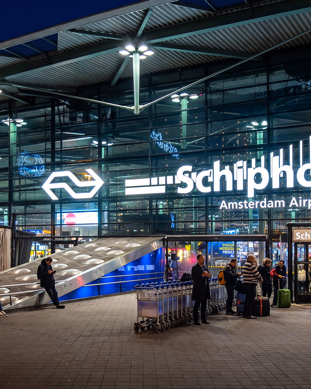 Amsterdam Airport Schiphol (AMS) terminal entrance illuminated at night with travellers and luggage trolleys